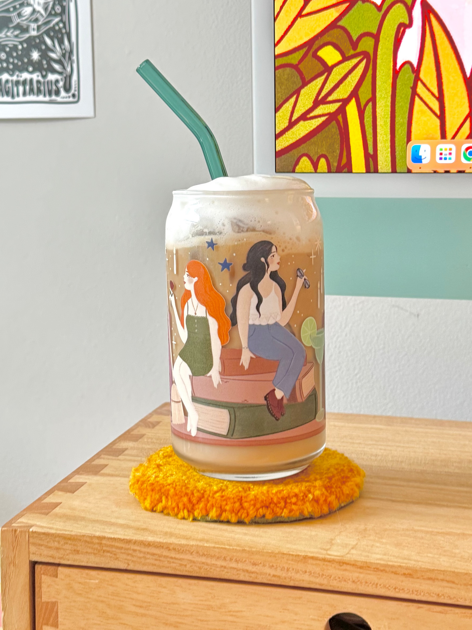 A clear glass can-shaped tumbler with a colorful illustrated print of of the owens sisters, witches from practical magic, filled with an iced coffee drink and topped with whipped cream, set on a yellow fuzzy coaster on a wooden table.