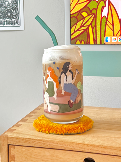 A clear glass can-shaped tumbler with a colorful illustrated print of of the owens sisters, witches from practical magic, filled with an iced coffee drink and topped with whipped cream, set on a yellow fuzzy coaster on a wooden table.