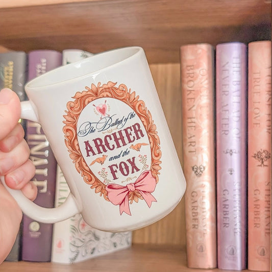 Mug with 'The Archer and the Fox' design held in front of a bookshelf with books.